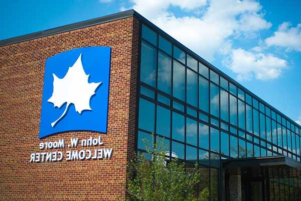 The John W. Moore Welcome Center is seen in the daytime, with blue sky and white clouds reflected in the building’s windows to the left. A large white Sycamore leaf on blue background is seen on the building’s red brick wall to the right, with the name underneath. 