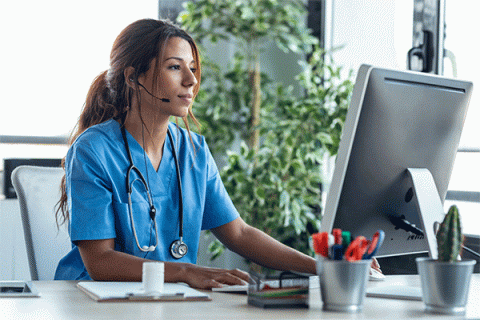 Female with long brown hair wearing blue nursing scrubs looking at a desktop computer screen while sitting at a table.  