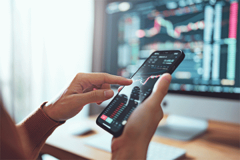 Detail image of person holding a phone with financial information on the screen with more information on a computer screen in the background. 